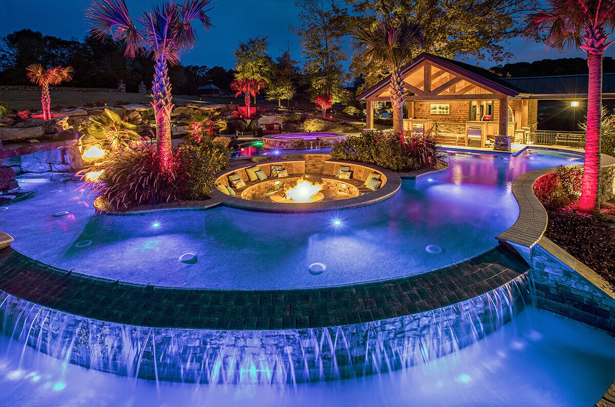 pogo of a backyard pool design in PebbleTec PebbleBrilliance Deep Cove Pool Finish medium blue water color with a fire pit in the center and waterfall features submitted for World's Greatest Pools 2021