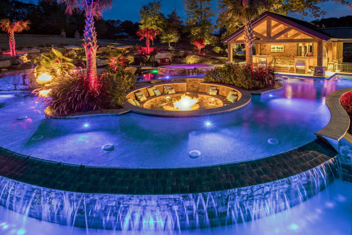 pogo of a backyard pool design in PebbleTec PebbleBrilliance Deep Cove Pool Finish medium blue water color with a fire pit in the center and waterfall features submitted for World's Greatest Pools 2021